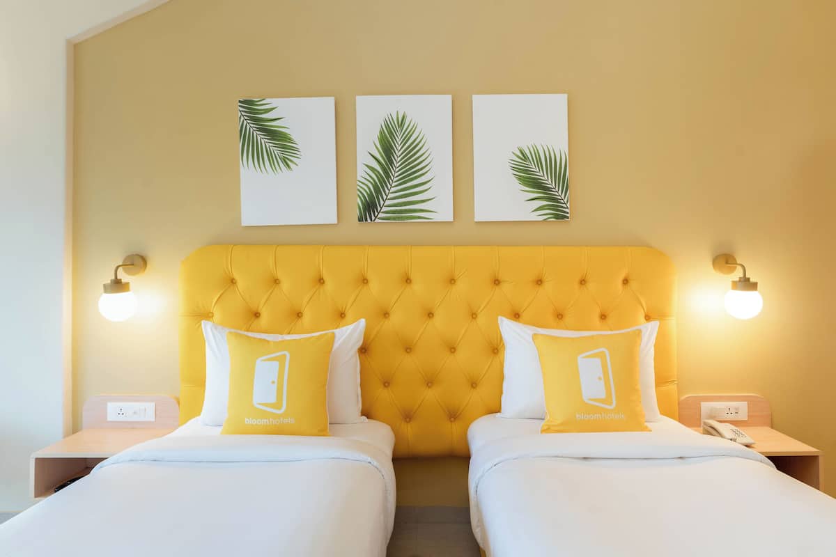 A hotel room with a yellow tufted headboard, two beds with white bedding and yellow pillows, a bedside table with a lamp, and a wall-mounted phone.