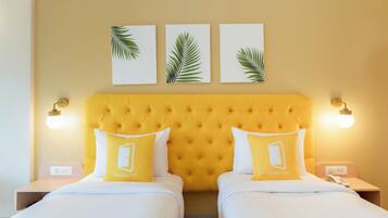 Habitación de hotel con una tapizada de color amarillo, dos camas con ropa de cama blanca y almohadas amarillas, una mesita de noche con lámpara y un teléfono en la pared.