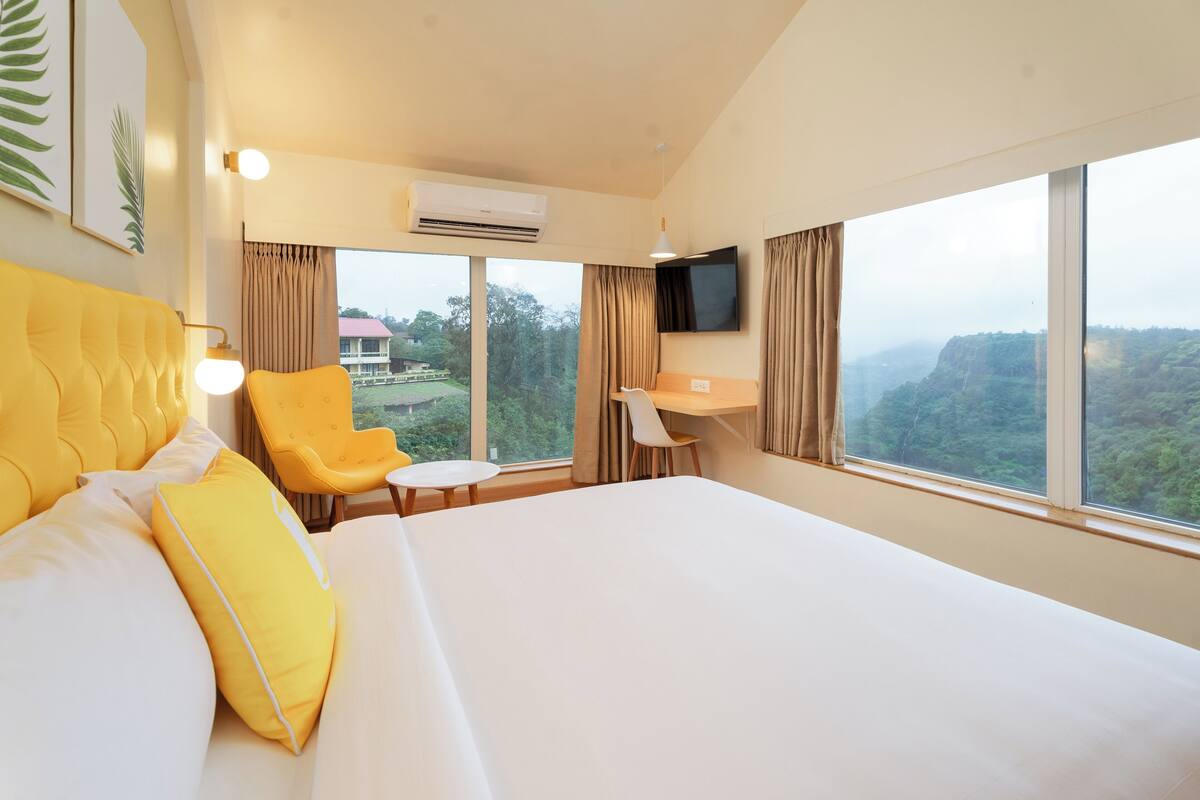 A hotel room with a large bed, a desk, and a chair. There is a TV mounted on the wall and a window with a view of the outdoors.