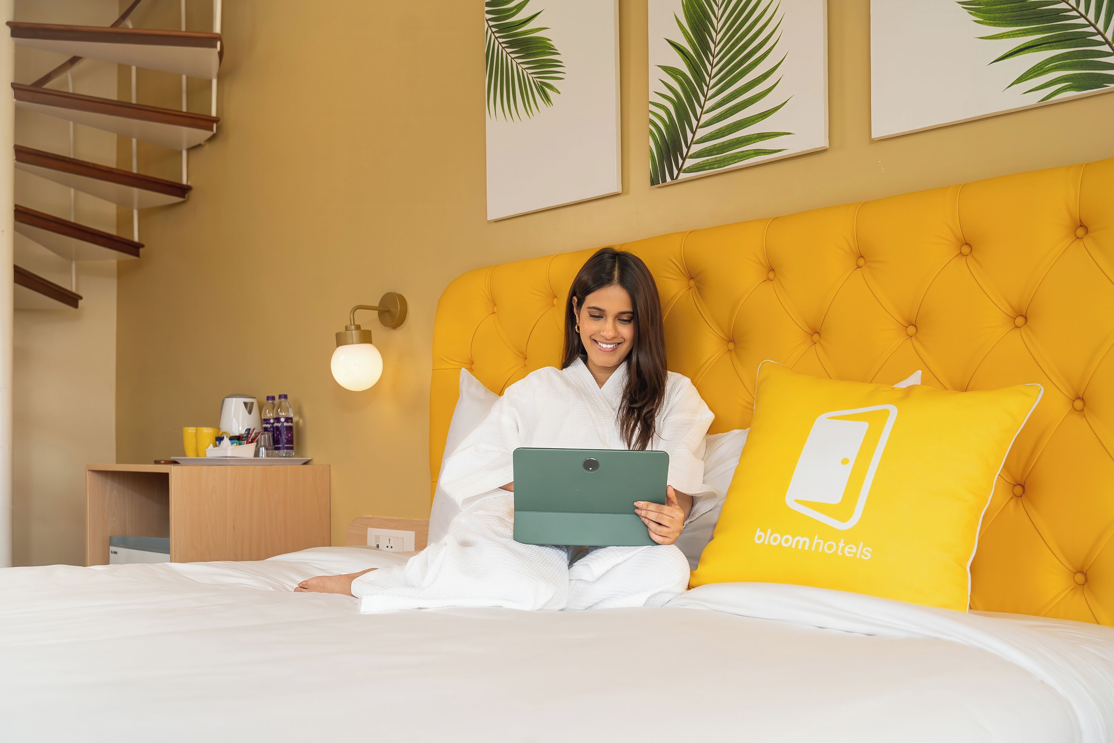A bed with a yellow headboard, a bedside table with a lamp, and a wall with framed leaves.