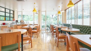 A modern cafe with round tables and chairs, a green cushioned bench, and large windows with a view of greenery. - Bloom Hotel Khandala (Mawal)