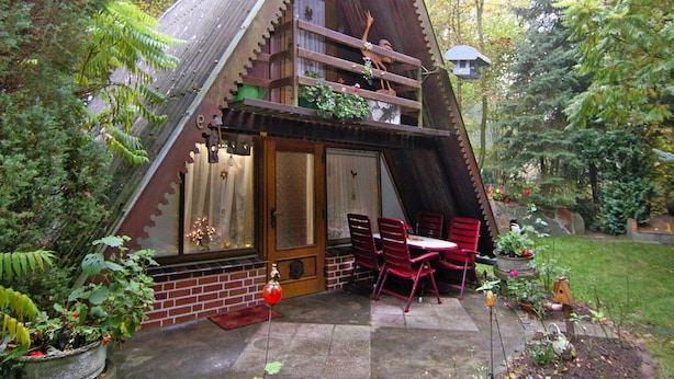 Une charmante maison en bois de forme triangulaire, dotée d’un balcon, d’un coin repas avec des chaises rouges et d’un jardin agrémenté de plantes en pot.