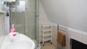 Un baño con un lavamanos blanco, un box de ducha de vidrio, un carrito de almacenamiento blanco y un perchero con una toalla marrón.