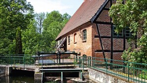 Un edificio storico in mattoni con tetto spiovente, un piccolo ponte su un canale, e rigogliosa vegetazione tutt'intorno.