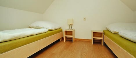 A small, triangular room with two wooden bunk beds, each with a green mattress and white pillows. There is a small wooden nightstand with a lamp between the beds.