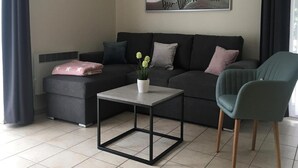 Ένα μοντέρνο σαλόνι με έναν γκρι καναπέ, ένα μικρό τραπεζάκι και μια πράσινη πολυθρόνα. Στον τοίχο κρέμεται ένας πίνακας που απεικονίζει μια παραθαλάσσια σκηνή.