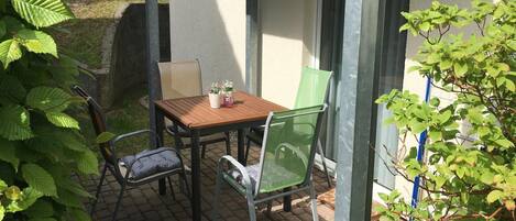 A patio with a wooden table and two chairs, surrounded by greenery.