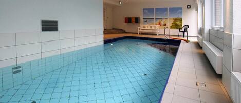 An indoor swimming pool with a tiled floor, a bench, and a view of the sea through the windows.