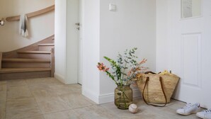Ein heller, minimalistischer Flur mit einer Holztreppe, einer Glasvase mit Blumen, einer gewebten Tasche und einem Baseball auf dem Boden.