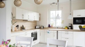 Eine moderne Küche mit weißen Schränken, einer Holzausziehtheke und einem eingebauten Backofen. Es gibt Pendelleuchten, ein Fenster, das für natürliches Licht sorgt, und eine Spüle mit Armatur.