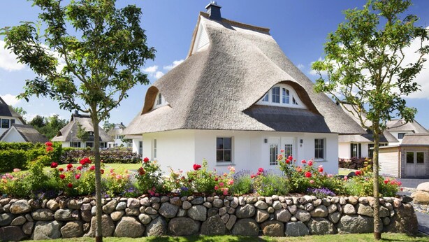Ein Haus mit einem Strohdach, einer Steinmauer und Blumenbeeten.