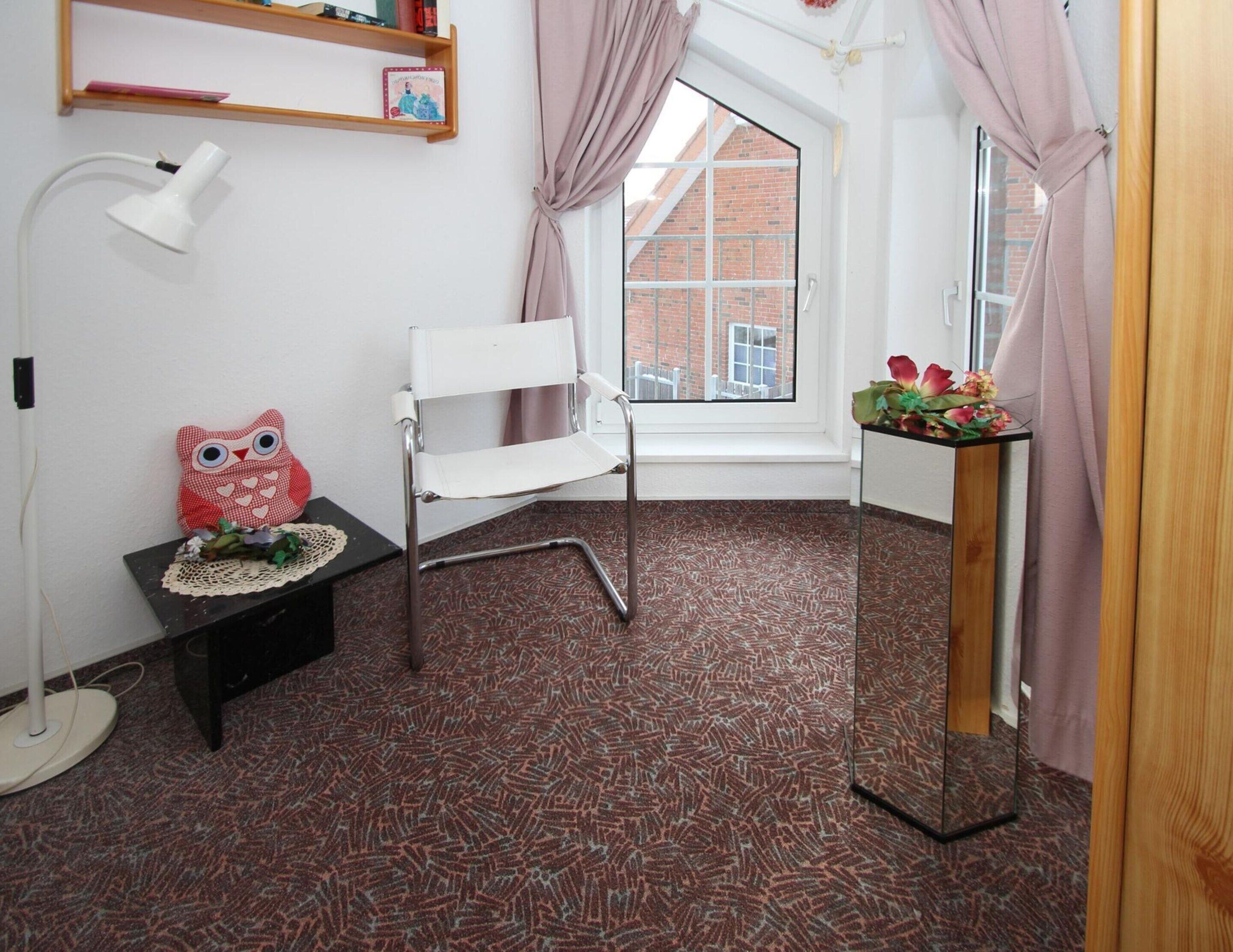 A small room with a white chair, a small table with a red owl decoration, a lamp, and a window with curtains.