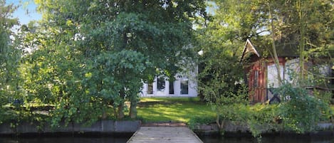 A wooden dock leading to a white building with a triangular roof, surrounded by lush greenery and a calm water body.
