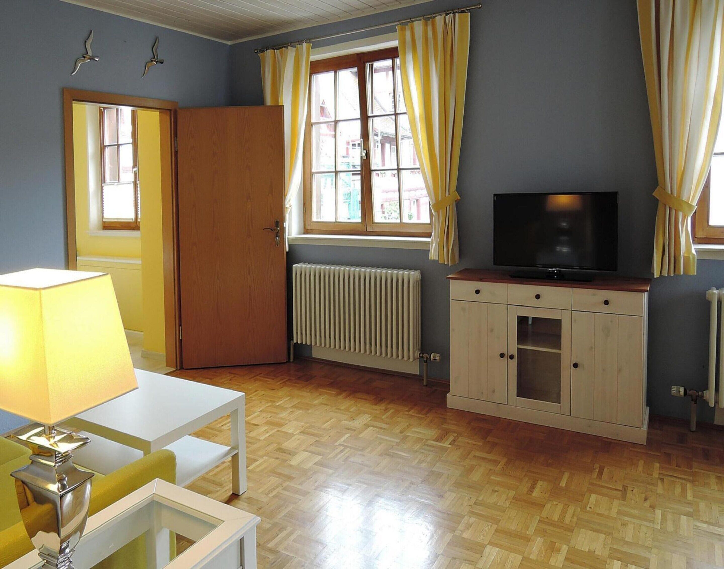 Une pièce avec un plancher en bois, une table blanche avec une lampe, un téléviseur posé sur un meuble en bois et une fenêtre avec des rideaux jaunes.