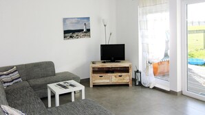 A living room with a grey sofa, a white coffee table, a wooden TV stand, and a painting of a lighthouse. - Starfish Holiday Home in Rerik Near Beach (Rerik)