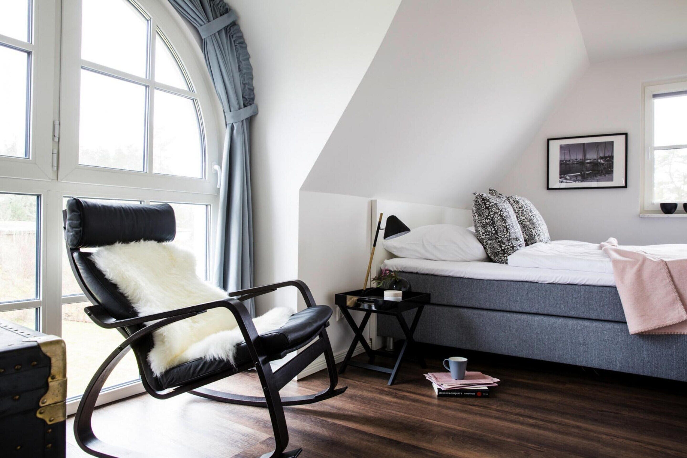 A bedroom with a bed, a black leather chair with a white fluffy pillow, a bedside table with a lamp, and a window with curtains.