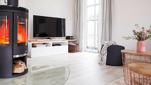 Ein modernes Wohnzimmer mit einem schwarzen Kaminofen, einem Flachbildfernseher, einer weißen Unterhaltungsecke, einem großen Fenster mit Vorhängen, einem runden Couchtisch und einem Blumentopf.