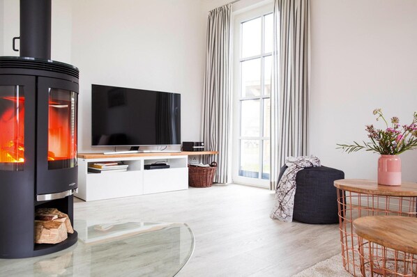 A modern living room with a black wood-burning stove, a flat-screen TV, a white entertainment unit, a large window with curtains, a round coffee table, and a potted plant. - Luxushaus in Bodstedt mit Sauna und Lagune (Fuhlendorf)