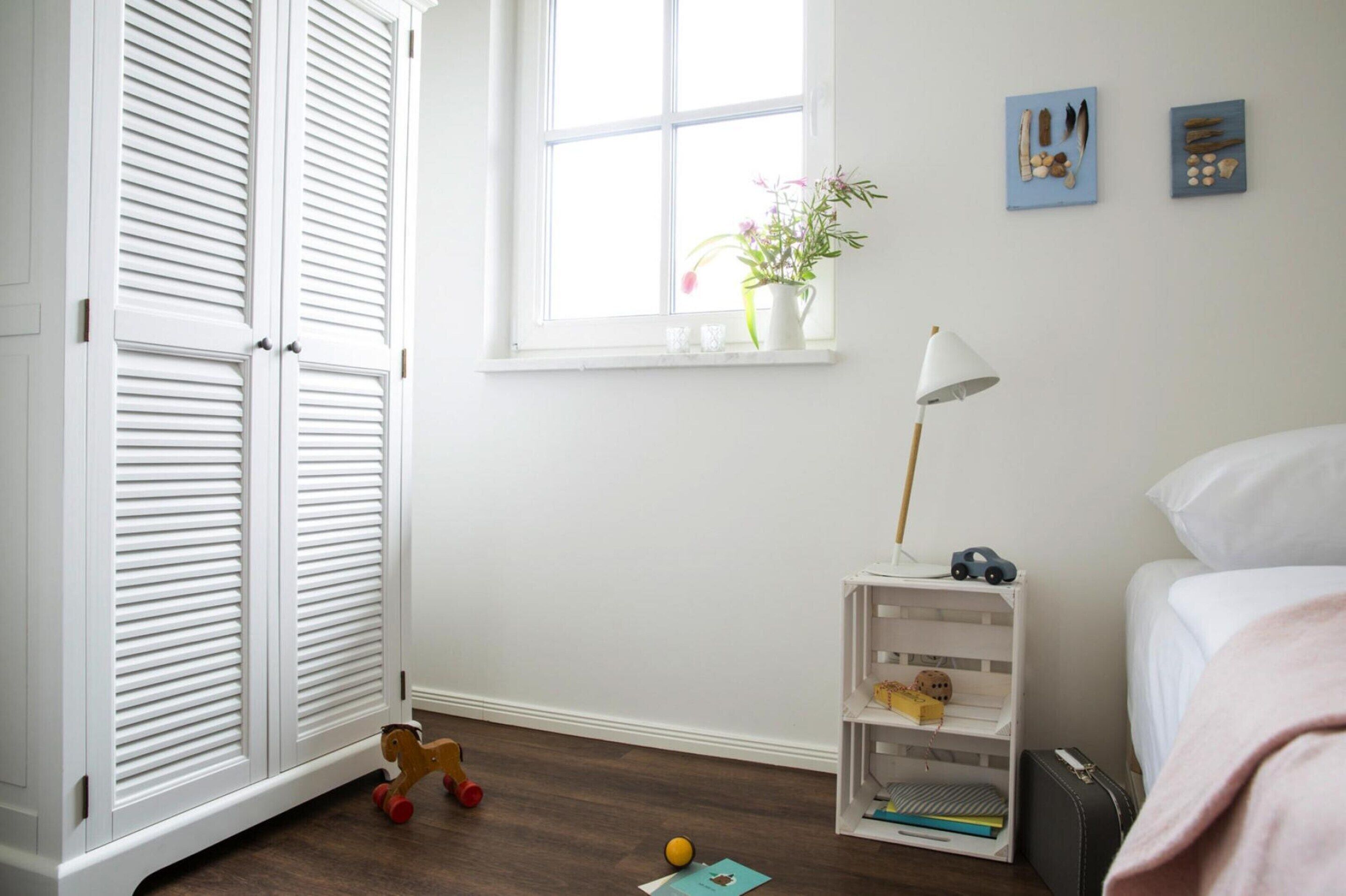 A child's room with a bed, a white wardrobe, a window with a plant, and a shelf with toys.