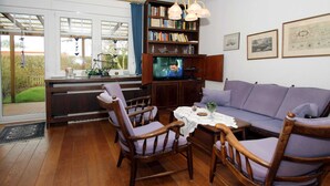 Een woonkamer met een houten vloer, een paarse bank, twee houten stoelen, een salontafel, een boekenkast en een televisie.
