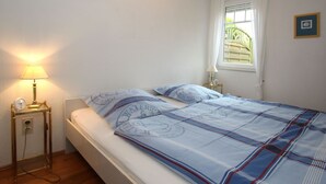 Een net opgemaakt bed met een blauw-wit geruite sprei en passende kussens, een nachtkastje met een lamp, en een raam met uitzicht op een groene brug.