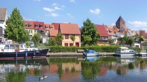 A calm river with boats docked on the left, surrounded by traditional European-style buildings with red roofs and a church tower. - Appartement Hafenflair am Plauer See (Plau am See)