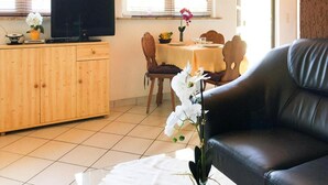 A living room with a black leather sofa, a wooden cabinet with a television, and a dining area with a table and chairs. - Reihenhaus am Kummerower See, Sommersdorf (Sommersdorf)