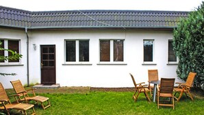 A white building with a tiled roof, a front door, and windows. There is a grassy lawn with wooden chairs and a table. - Reihenhaus am Kummerower See, Sommersdorf (Sommersdorf)