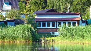 Ein Haus mit rotem Dach und weißen Wänden, umgeben von hohem Gras und einem Gewässer.