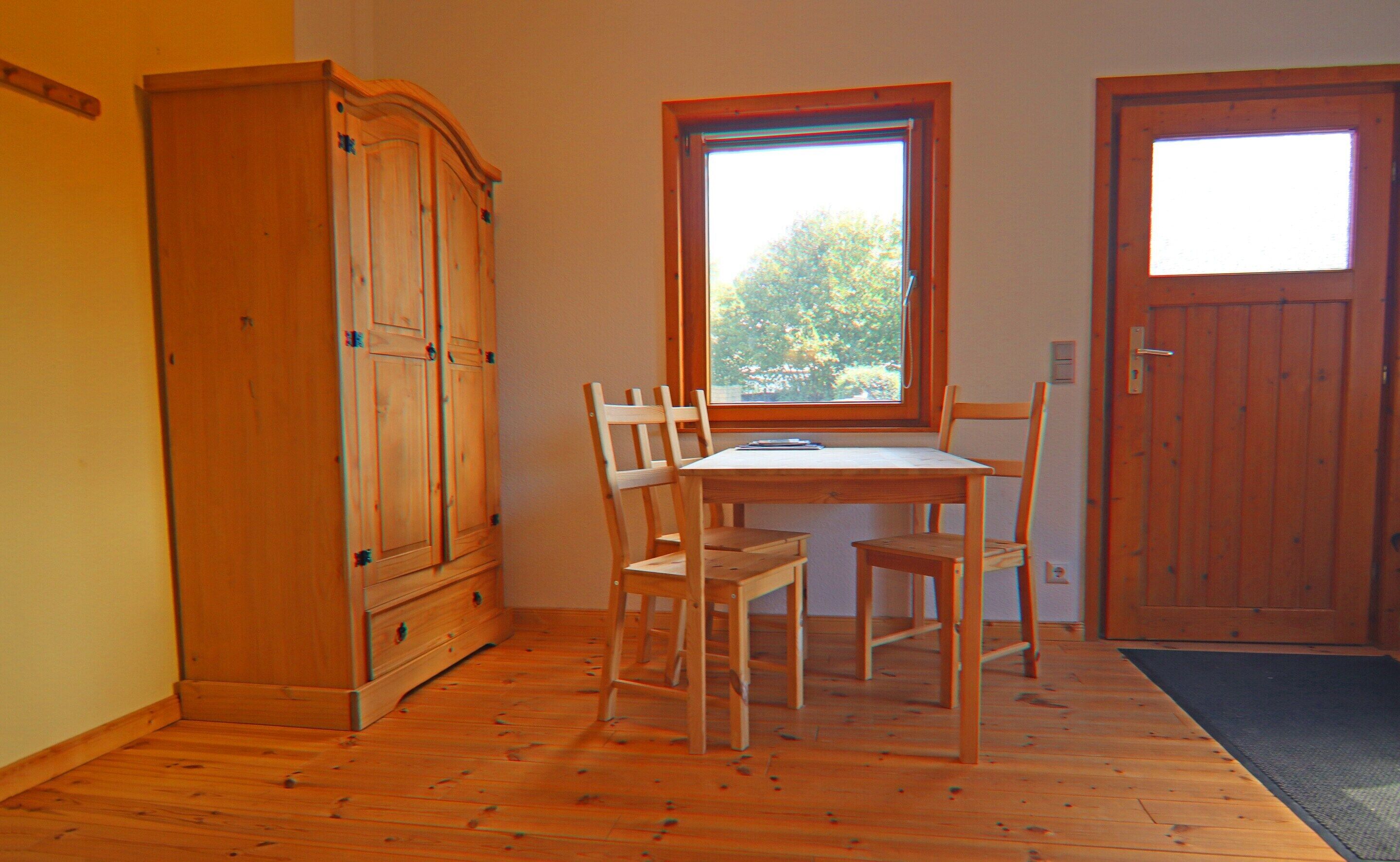 A room with a wooden cabinet, a table with chairs, a window with a view of trees, and a door.