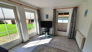 A room with a large sliding glass door leading to a garden, a flat-screen TV, a small table, and a patterned rug. - Ferienanlage Petersdorf auf Fehmarn-petersdorf (Fehmarn)