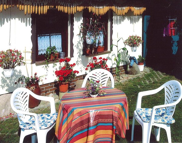 An outdoor patio with a table and chairs, surrounded by potted plants and a white wall with windows.