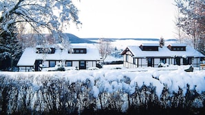 A snow-covered landscape with two buildings, one with a gabled roof and another with a flat roof, surrounded by trees and a distant view of hills. - Feriendorf Ofingen, Bad Durrheim (Bad Duerrheim)