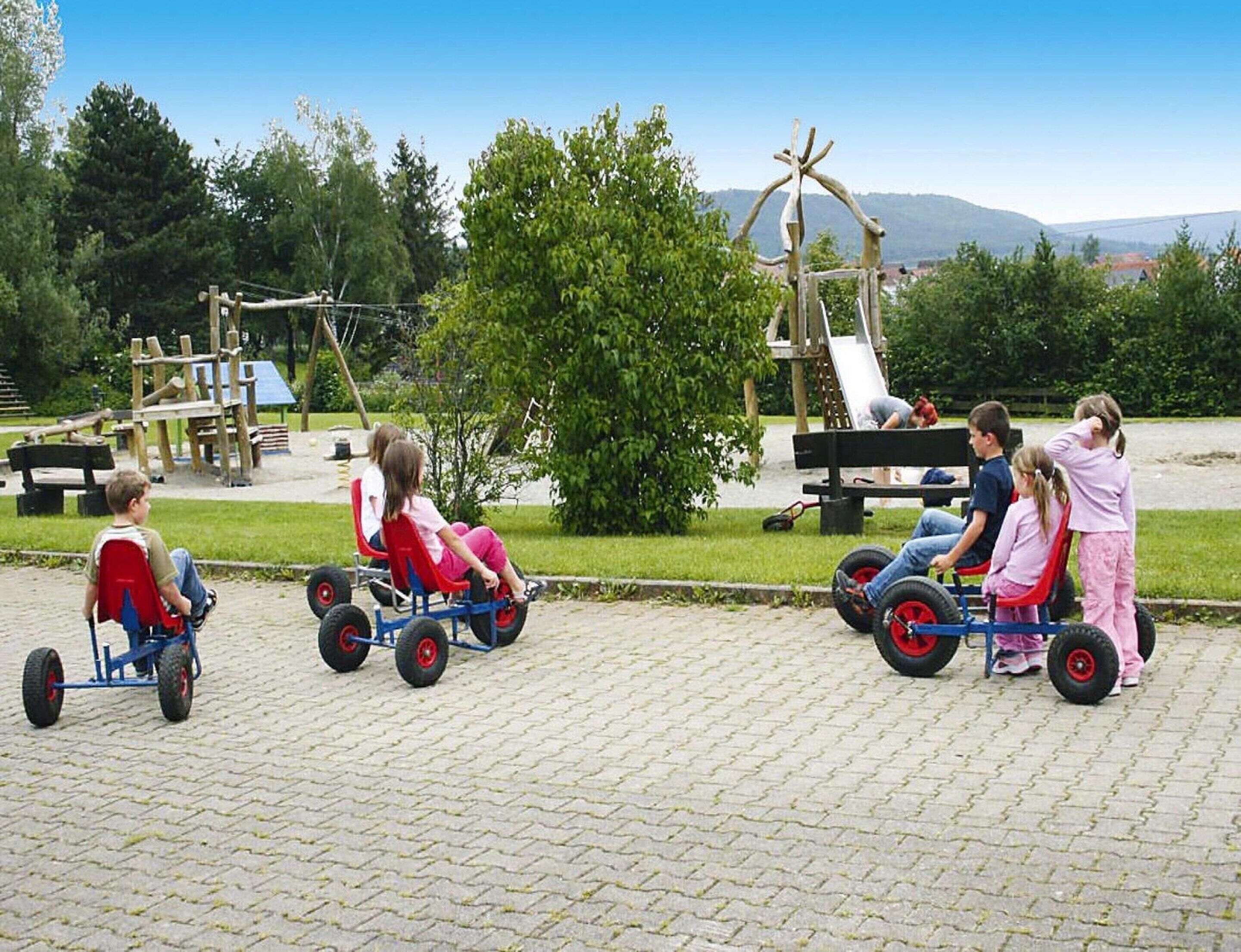 Niños montando triciclos por un sendero pavimentado en un parque con juegos infantiles y árboles al fondo.