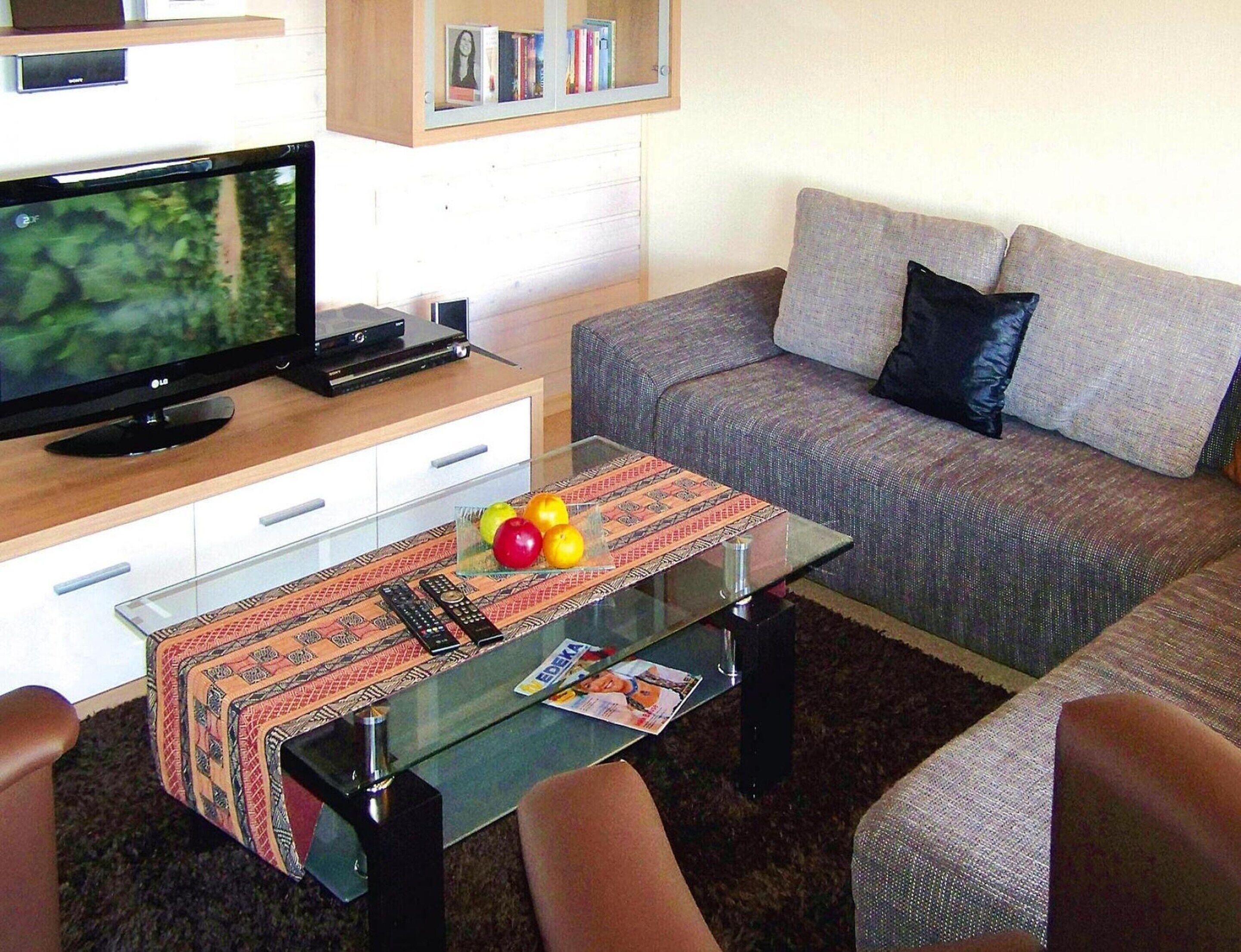 A living room with a flat-screen TV, a glass coffee table with fruit, a sofa, and a bookshelf.