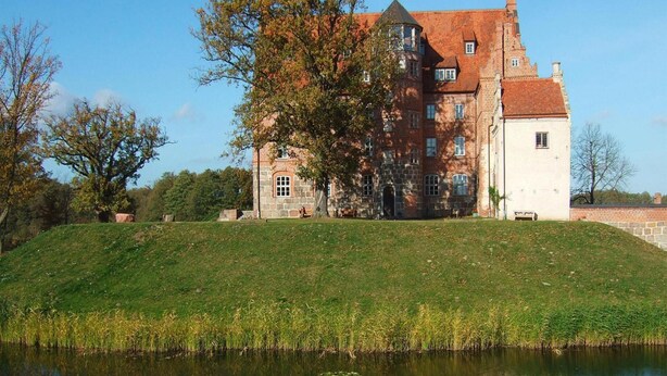 A large, historic building with multiple windows and a tower, situated on a grassy hill with a body of water in the foreground. - Ferienwohnungen am Flacher See, Klocksin (Klocksin)