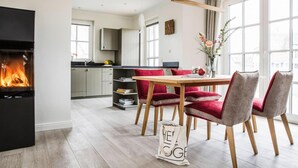 A modern kitchen with a fireplace, a dining area with red chairs, and a view of the city through large windows. - Ferienhaus, Fuhlendorf (Fuhlendorf)
