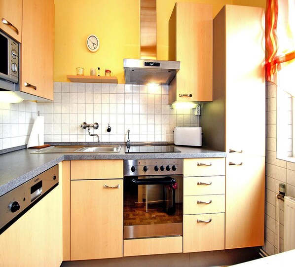 A kitchen with wooden cabinets, a stainless steel oven, a microwave, and a clock on the wall. - Bungalow Klonsnack in Dornumersiel (Dornum)