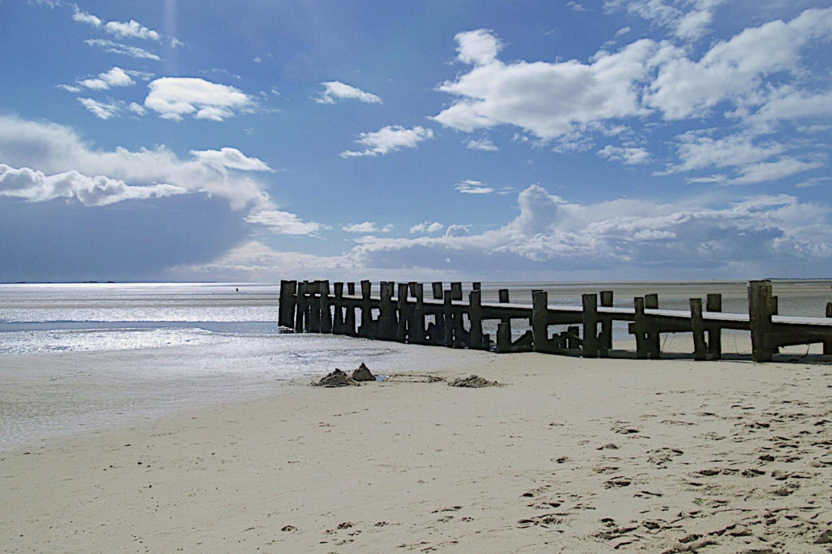 Une plage avec une jetée en bois qui s’avance dans l’eau, un rivage sablonneux et un ciel dégagé parsemé de nuages.