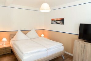 Una cama bien hecha con ropa blanca, una lámpara de noche, un televisor sobre un mueble de madera y un cuadro en la pared.