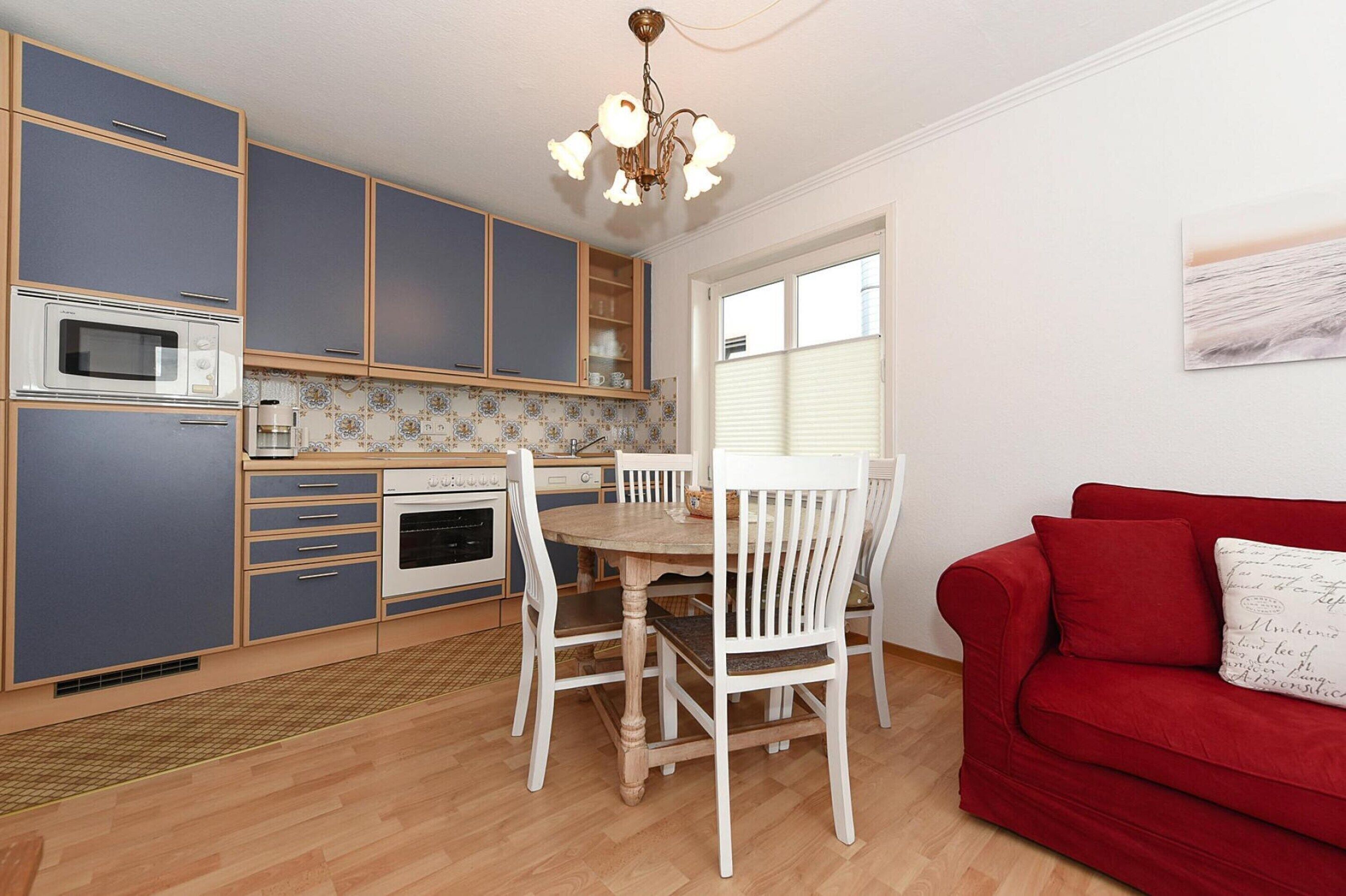 A compact kitchen with a dining table and chairs, a red sofa, and a window with blinds.