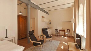 Une pièce spacieuse avec un grand lit, deux fauteuils, une petite table et un téléviseur posé sur un meuble en bois. On y trouve également un coin cuisine avec un évier et des placards, ainsi qu’une fenêtre qui laisse entrer la lumière naturelle.
