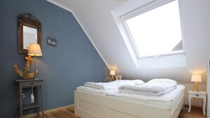 Ein Schlafzimmer mit schrägen Wänden, einem Oberlicht, einem Bett mit weißer Bettwäsche, zwei Nachttischen mit Lampen, einem kleinen Regal mit einer Lampe und einem wandmontierten Spiegel.