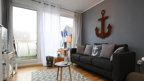 Ein Wohnzimmer mit einem dunkelgrauen Sofa, einem Teppich mit geometrischem Muster, einem Holztisch und einer großen Anker-Dekoration an der Wand.