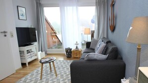Ein modernes Wohnzimmer mit einer grauen Couch, einem Fernseher auf einem weißen Schrank, einem gemusterten Teppich und Blick auf eine Treppe, die zu einem Balkon führt.