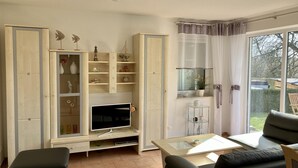 A living room with a TV, a glass coffee table, and a sofa. There is a window with curtains and a view of the outside.