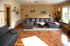 A living room with a sectional sofa, a coffee table, and a patterned rug. There are two wall-mounted speakers and a window with plants. - Ferienhaus Mowe in Mirow (Mirow)