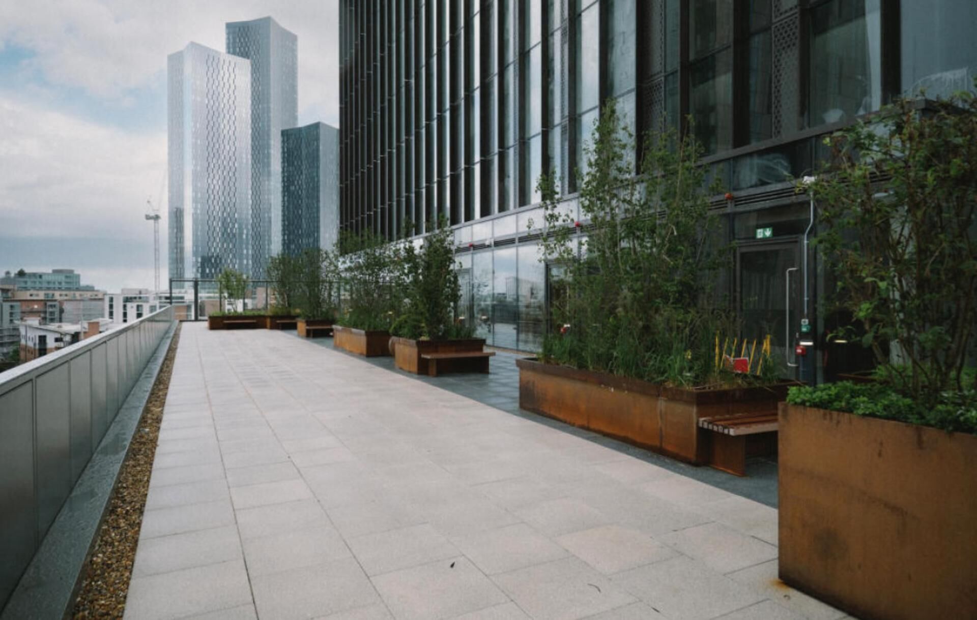 A rooftop terrace with a view of modern buildings, featuring a long, flat walkway and several planters with greenery.