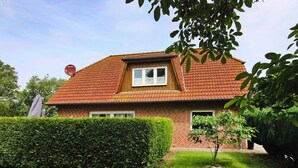 A two-story house with a red-tiled roof, a small window with white shutters, and a well-maintained hedge in the front yard. - Ferienwohnung im Ferienhaus Rosi, Fehmarn-schlagsdorf (Fehmarn)