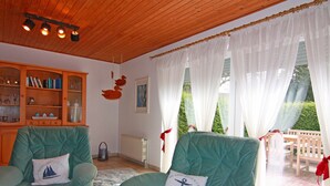A cozy living room with two green armchairs, a wooden bookshelf, and a view of the garden through large windows. - Ferienwohnung im Ferienhaus Rosi, Fehmarn-schlagsdorf (Fehmarn)